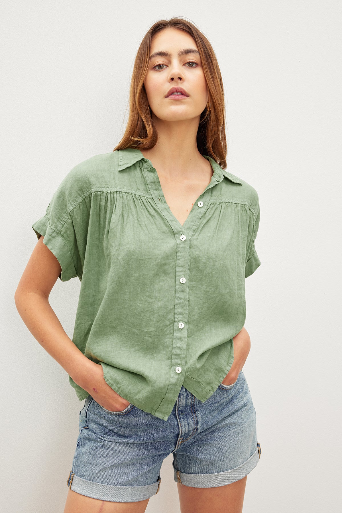 The model is wearing a Velvet by Graham & Spencer ARIA LINEN BUTTON FRONT TOP in sage green and denim shorts, exuding timeless appeal.-35955414991041