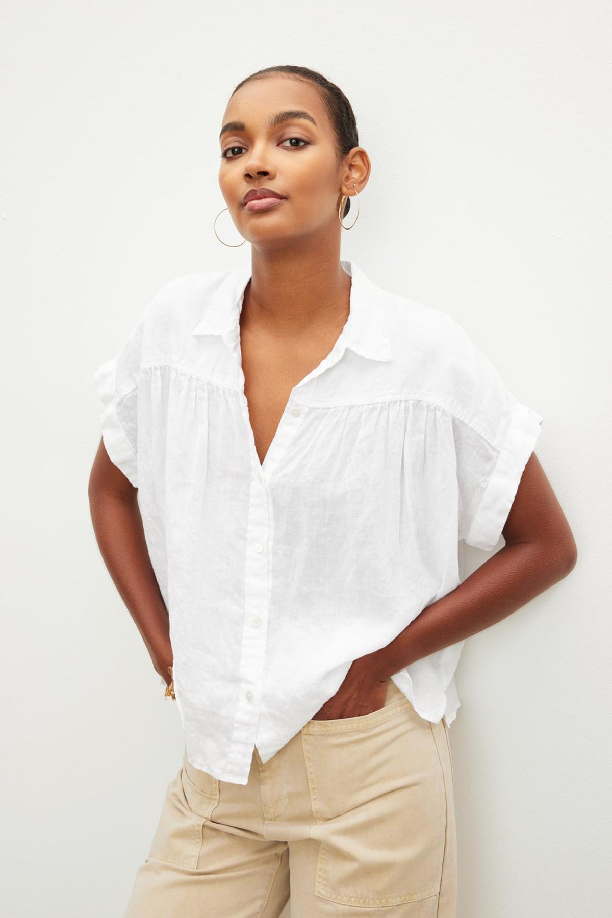   A confident woman posing in a Velvet by Graham & Spencer ARIA LINEN BUTTON FRONT TOP and beige pants against a plain background. 