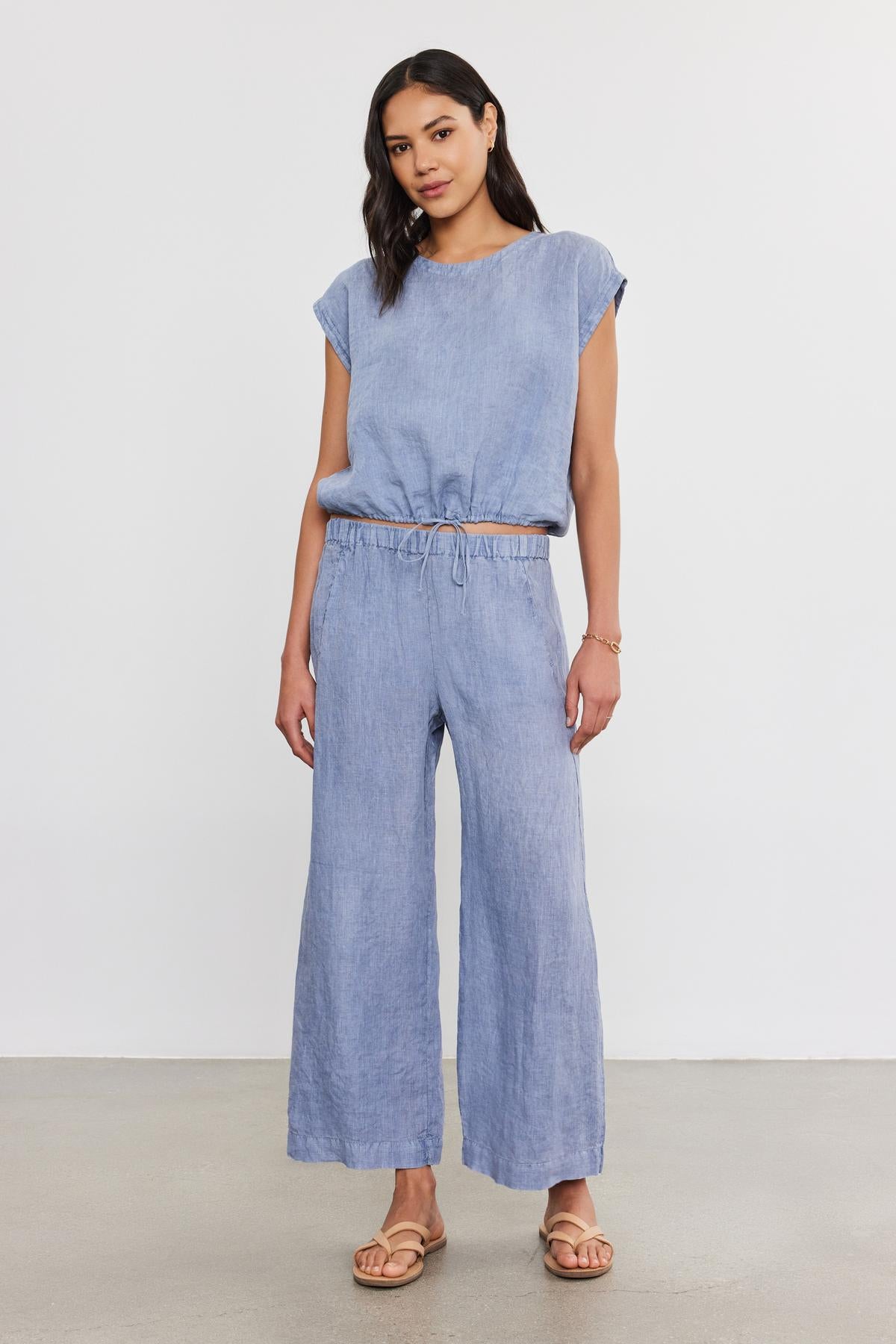 A woman in a blue denim jumpsuit and tan sandals with an ankle crop poses against a plain background.-36910133018817