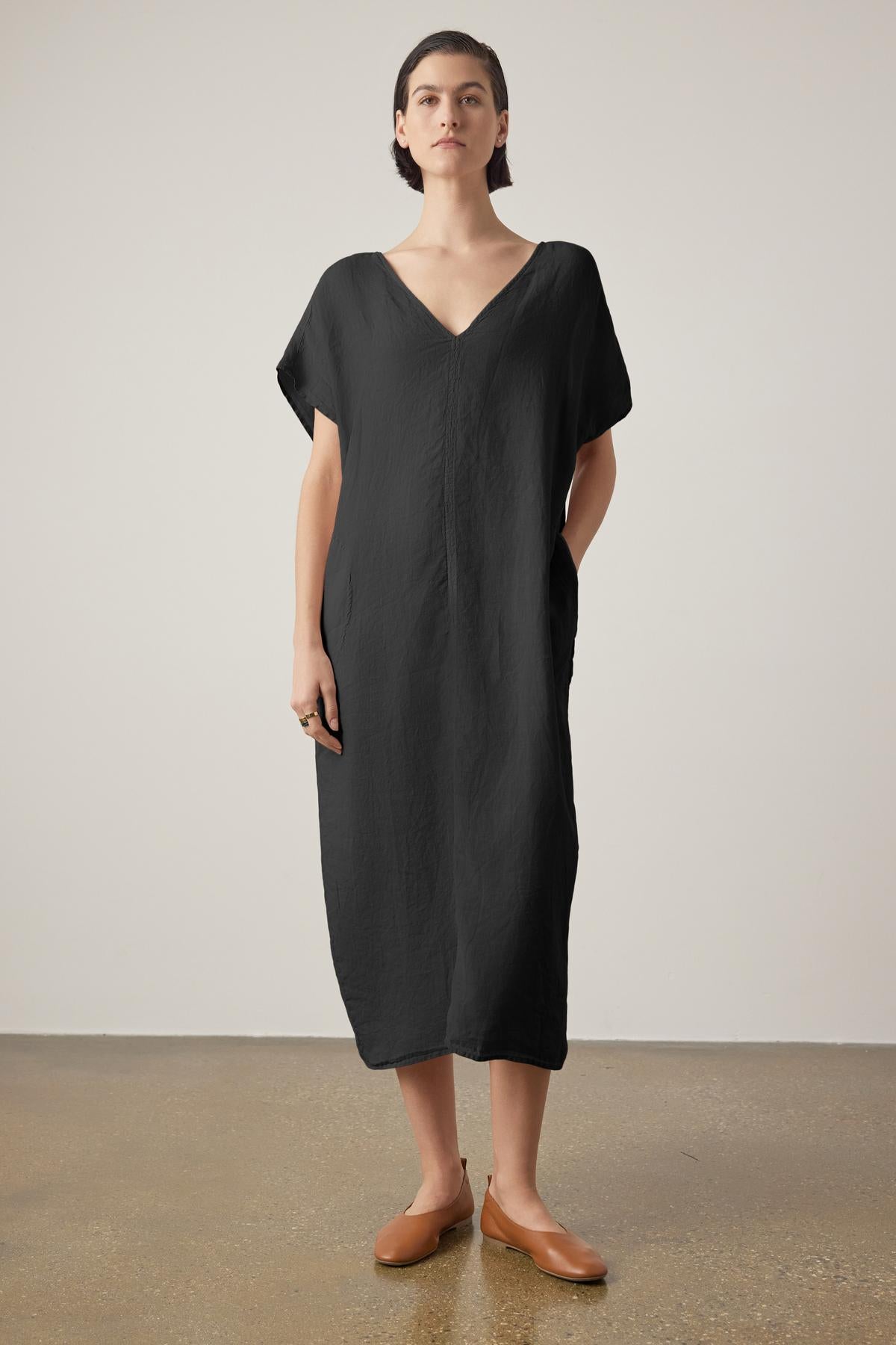 A woman in a loose black Montana linen dress by Velvet by Jenny Graham with a V neckline and brown loafers stands against a neutral backdrop, looking directly at the camera.-36863315345601