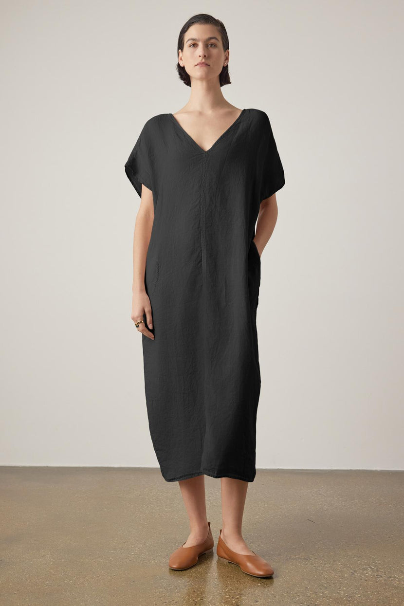 A woman in a loose black Montana linen dress by Velvet by Jenny Graham with a V neckline and brown loafers stands against a neutral backdrop, looking directly at the camera.
