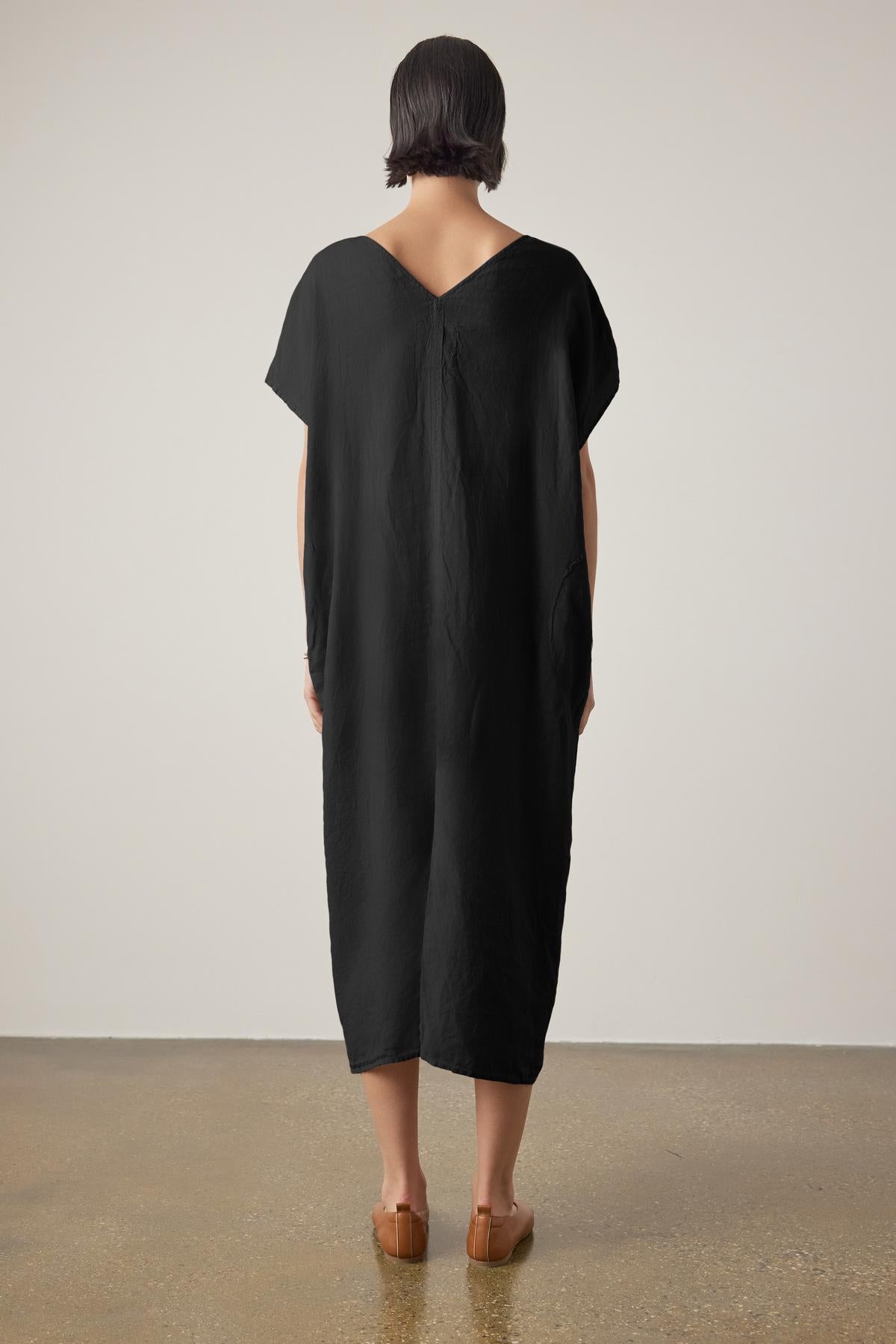 Woman standing with her back to the camera, wearing a black, loose-fitting Velvet by Jenny Graham Montana linen dress and brown heels on a neutral background.-36863315411137