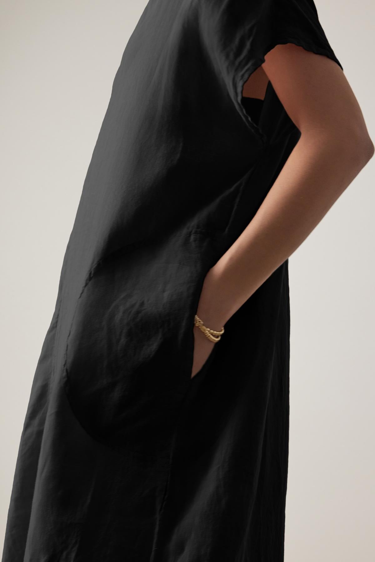 A close-up of a person wearing a Velvet by Jenny Graham black Montana linen dress with a cut-out detail at the elbow and a gold bracelet on the wrist.-36863315378369