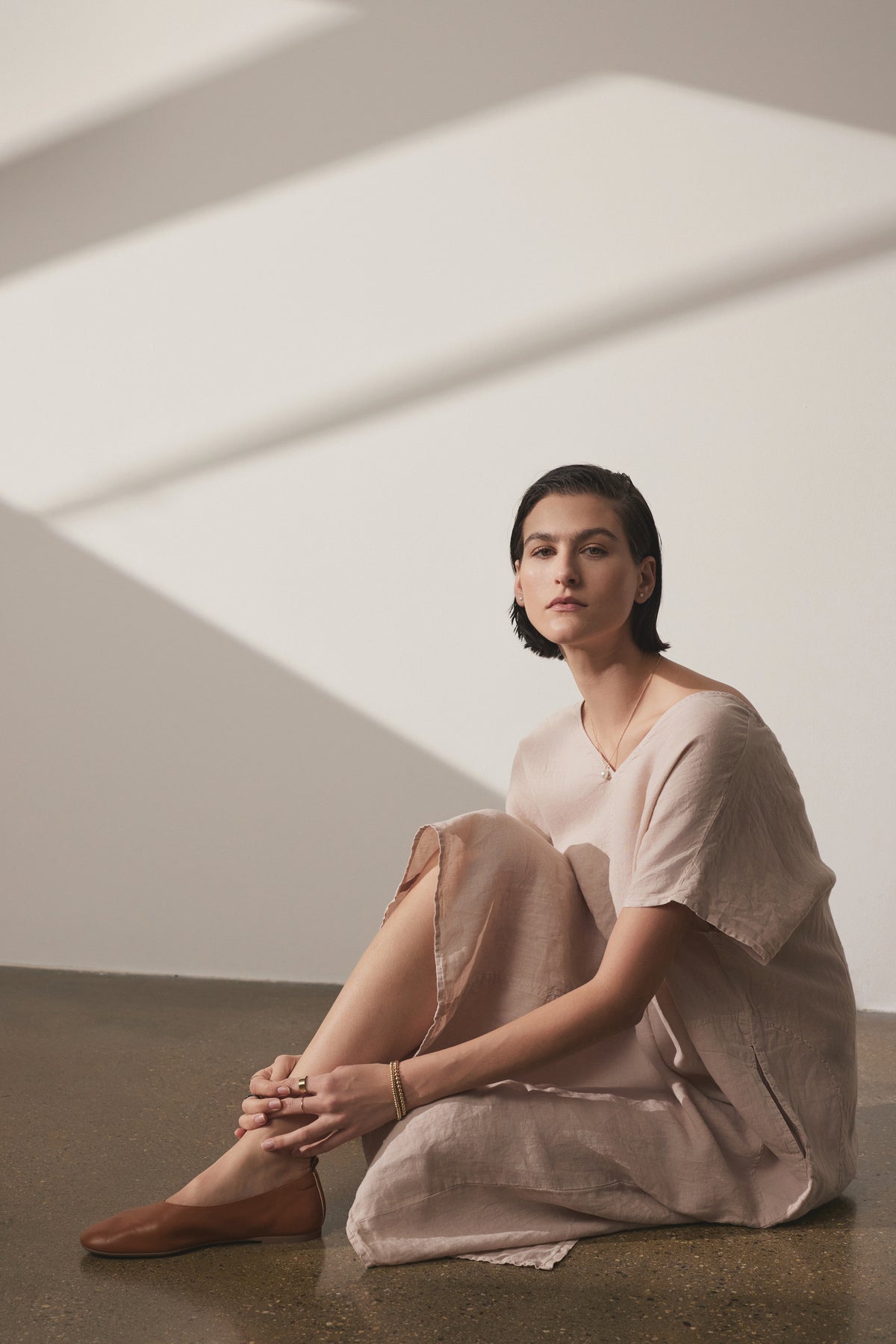 A woman in a Velvet by Jenny Graham Montana Linen Dress sits on the floor, leaning against a wall with diagonal light stripes, looking contemplatively towards the camera.-36863315181761