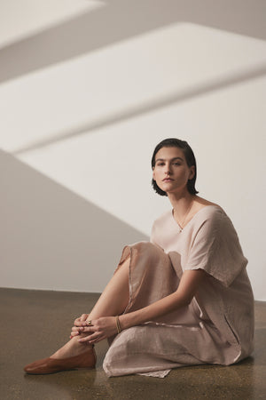 A woman in a Velvet by Jenny Graham Montana Linen Dress sits on the floor, leaning against a wall with diagonal light stripes, looking contemplatively towards the camera.