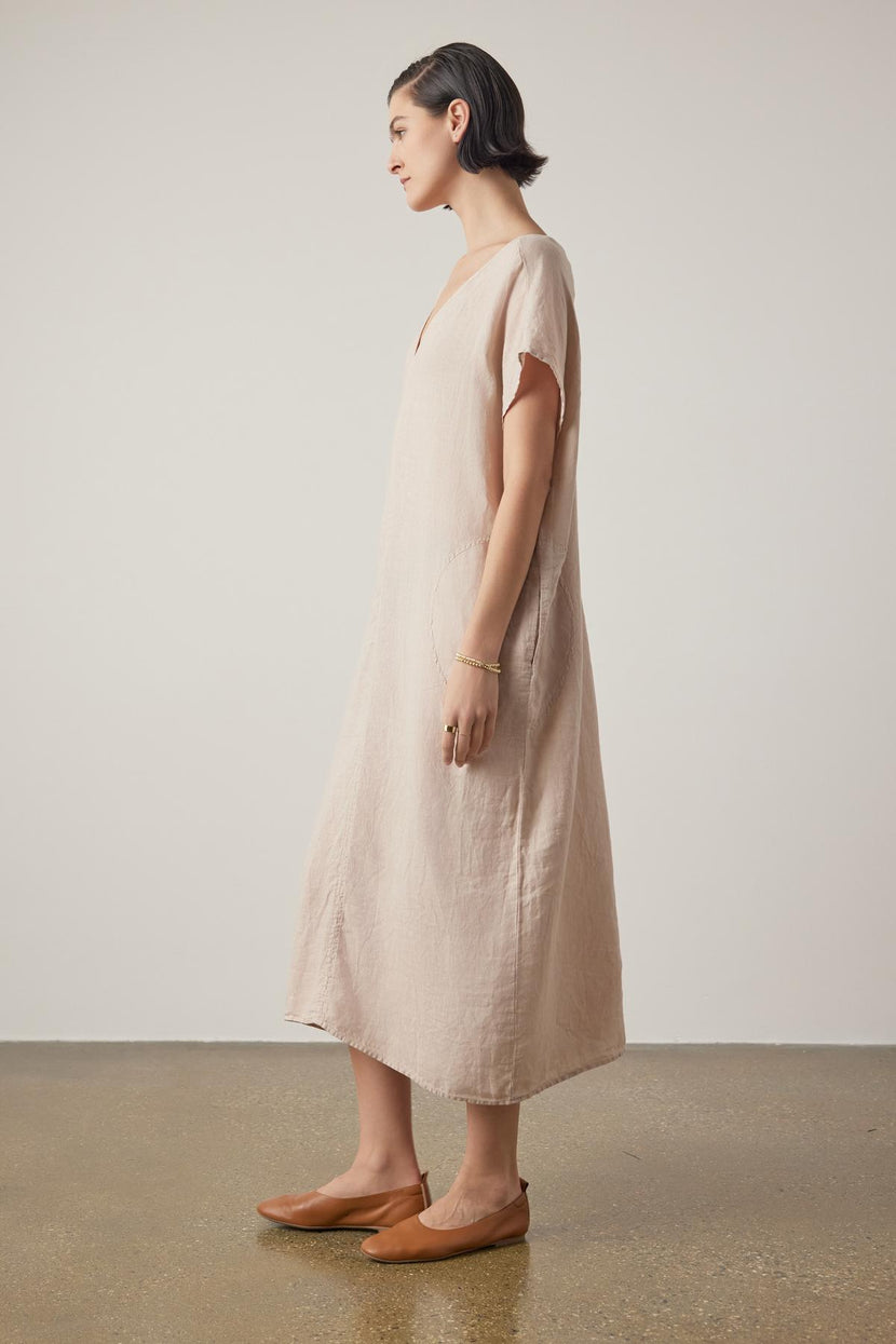 A woman in a Velvet by Jenny Graham Montana linen dress and brown loafers stands profile to the camera on a neutral background.