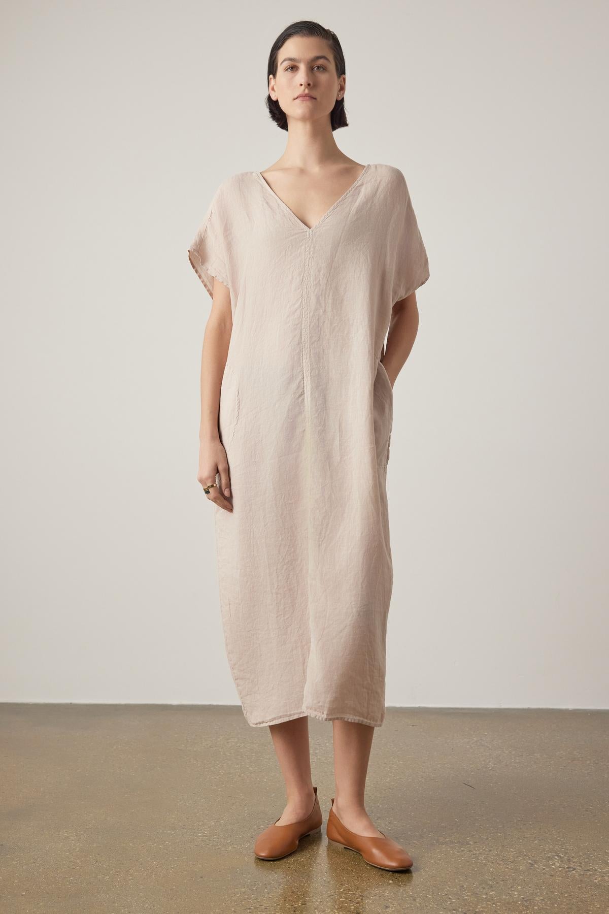 A woman stands facing forward, wearing a Velvet by Jenny Graham Montana linen dress with a V neckline and brown slip-on shoes, against a neutral backdrop.-36863315214529
