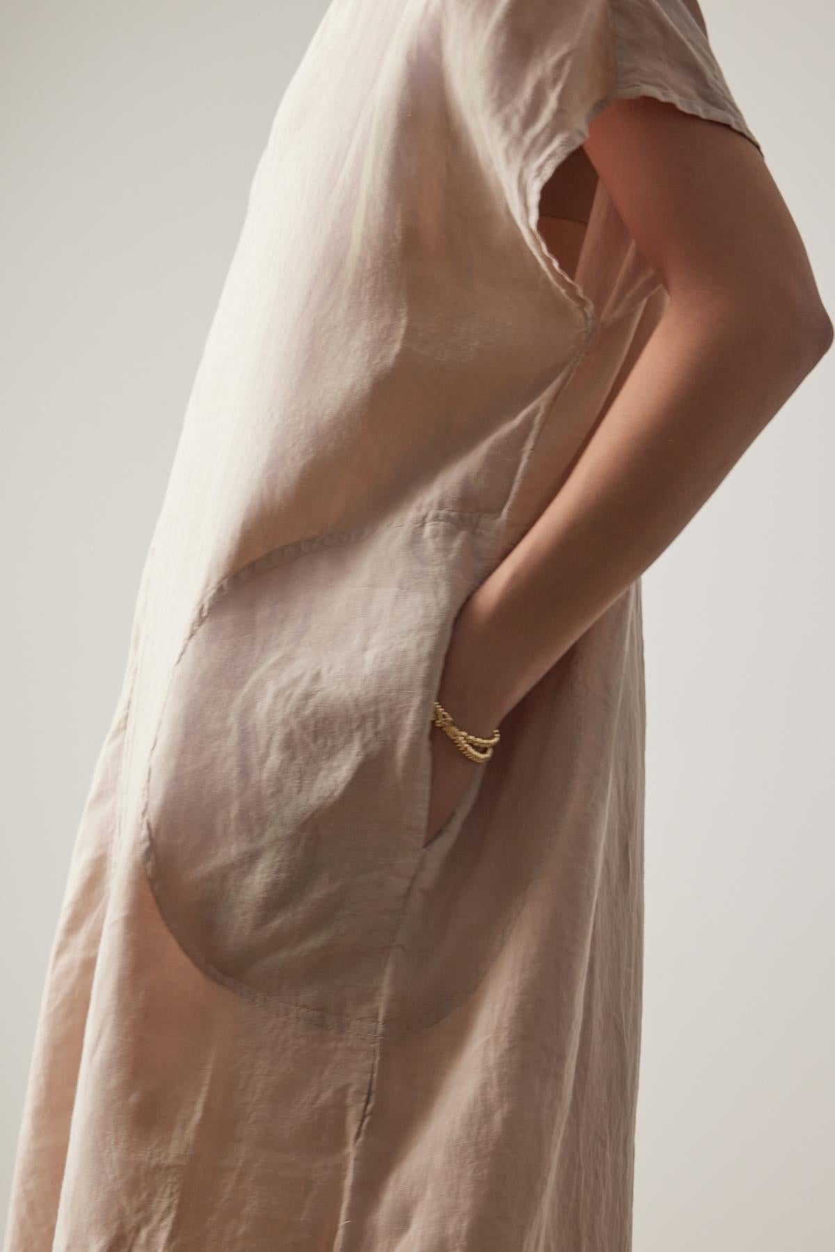 A close-up of a person in a Velvet by Jenny Graham Montana linen dress with a large pocket, highlighting a detail of a gold bracelet on their arm.-36863315312833