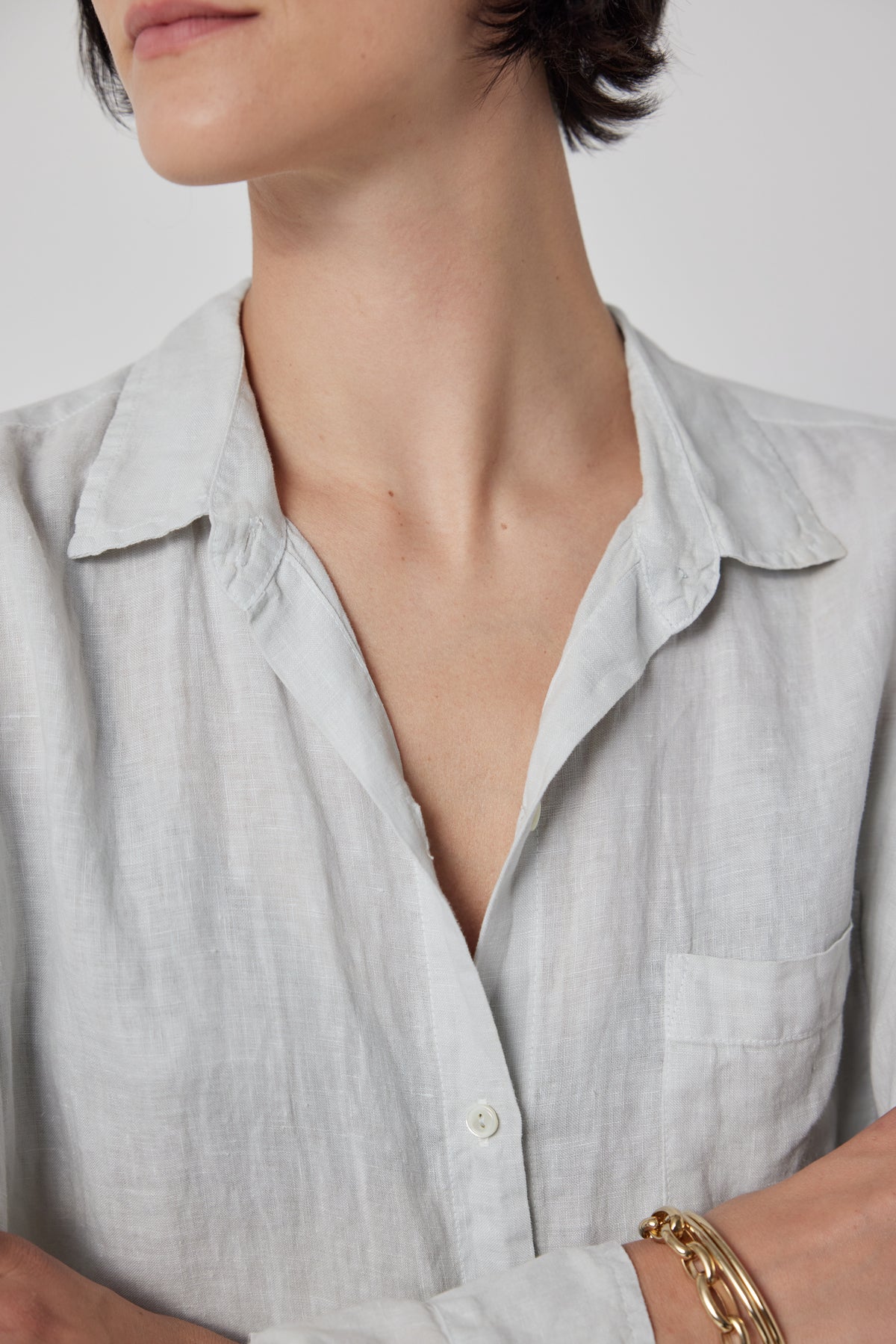   Close-up of a person wearing the Velvet by Jenny Graham MULHOLLAND LINEN SHIRT with a chest pocket. The person has short, dark hair and is accessorizing with a gold bracelet. The relaxed silhouette of the shirt features a scooped hemline. The background is plain white. 