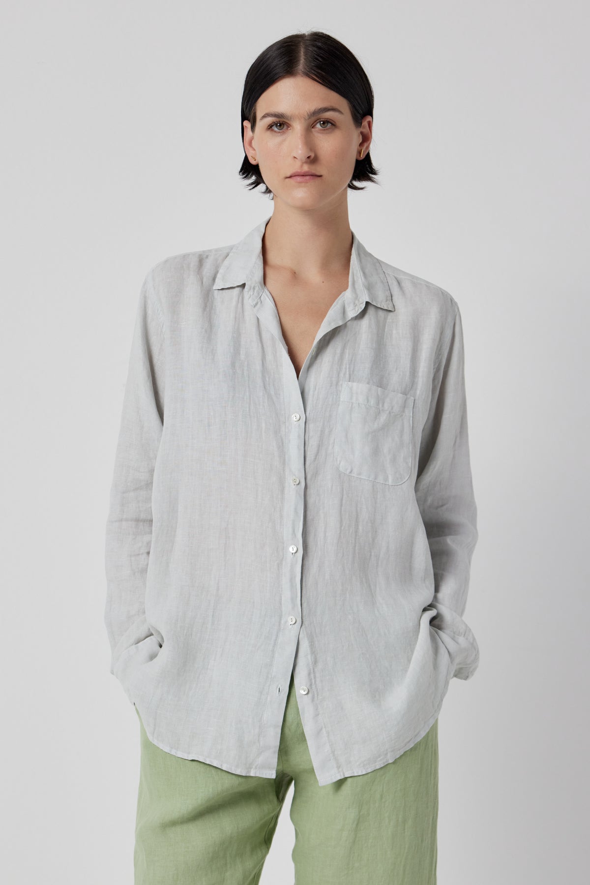   A person with short dark hair wearing a Velvet by Jenny Graham MULHOLLAND LINEN SHIRT in light gray with a scooped hemline and light green pants stands against a plain white background. 