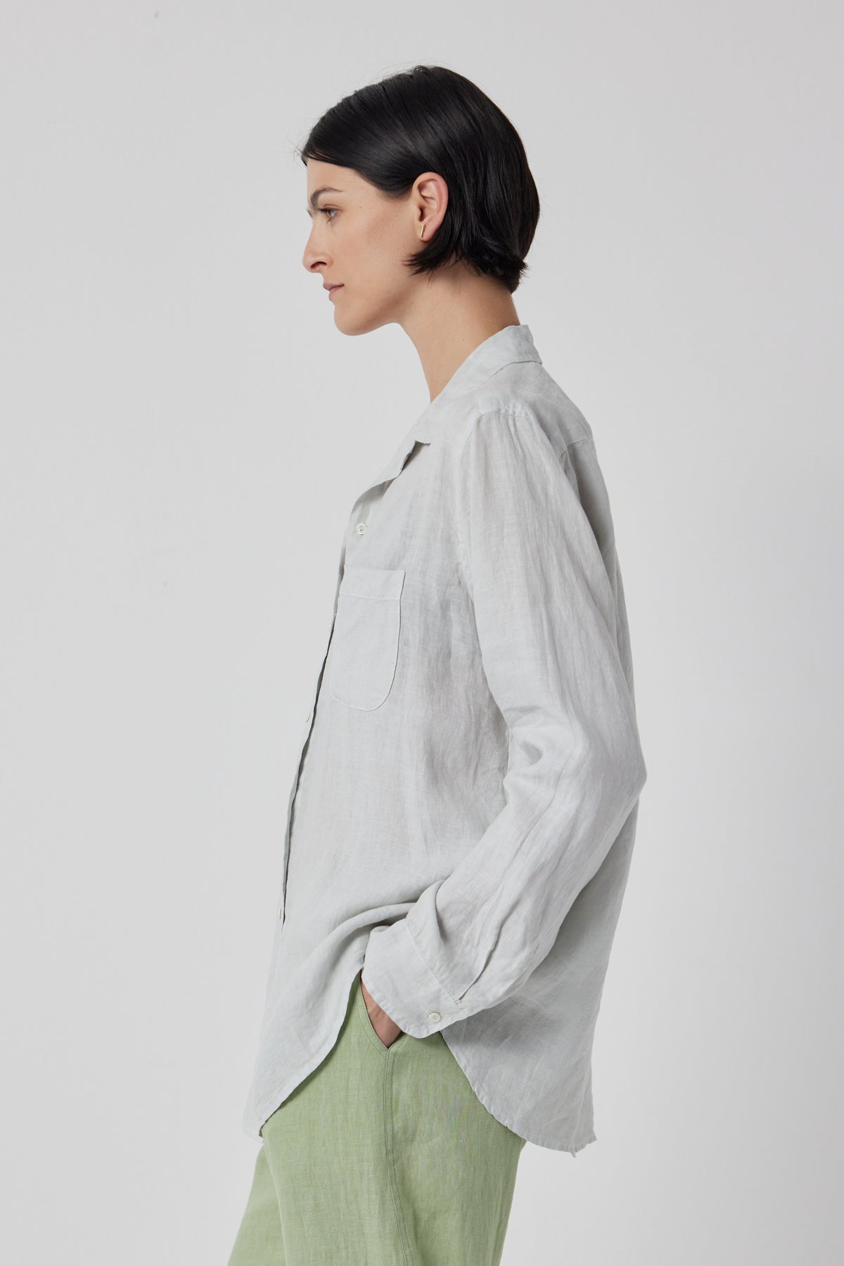 A person with short dark hair is standing in profile, wearing a MULHOLLAND LINEN SHIRT by Velvet by Jenny Graham with a scooped hemline and light green pants.-36212529725633