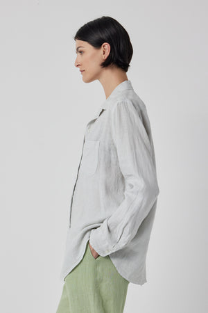 A person with short dark hair is standing in profile, wearing a MULHOLLAND LINEN SHIRT by Velvet by Jenny Graham with a scooped hemline and light green pants.