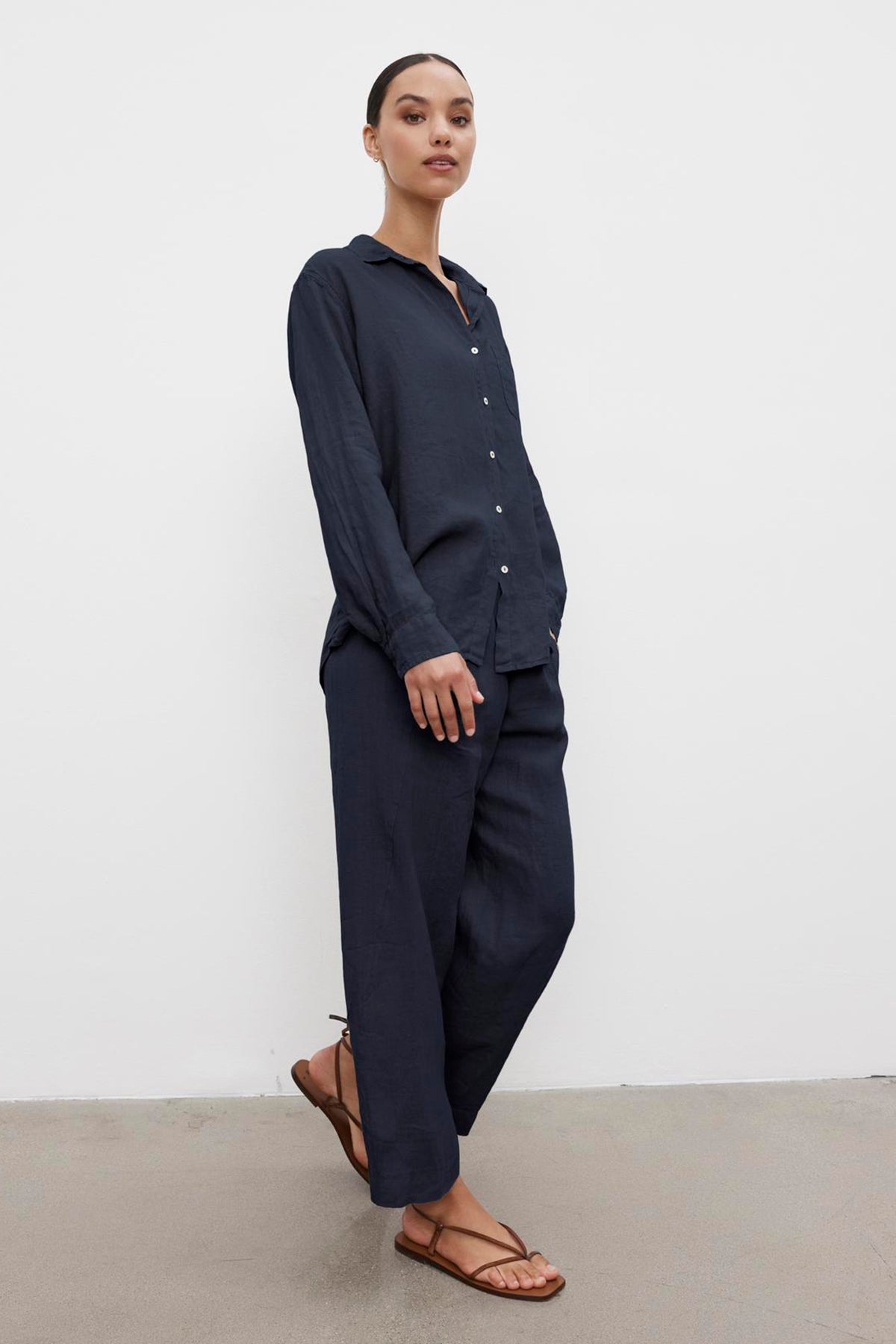 A person standing against a white background, wearing a dark blue MULHOLLAND LINEN SHIRT by Velvet by Jenny Graham with a relaxed silhouette, matching pants, and brown sandals.-36592968466625