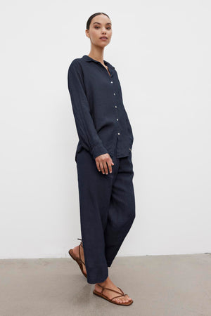 A person standing against a white background, wearing a dark blue MULHOLLAND LINEN SHIRT by Velvet by Jenny Graham with a relaxed silhouette, matching pants, and brown sandals.