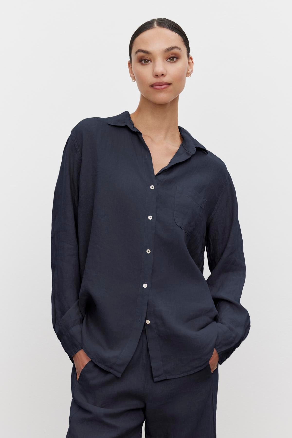 A person with dark hair tied back, wearing a dark blue MULHOLLAND LINEN SHIRT by Velvet by Jenny Graham and matching pants with a relaxed silhouette, stands against a plain white background with hands in pockets.-36592968401089