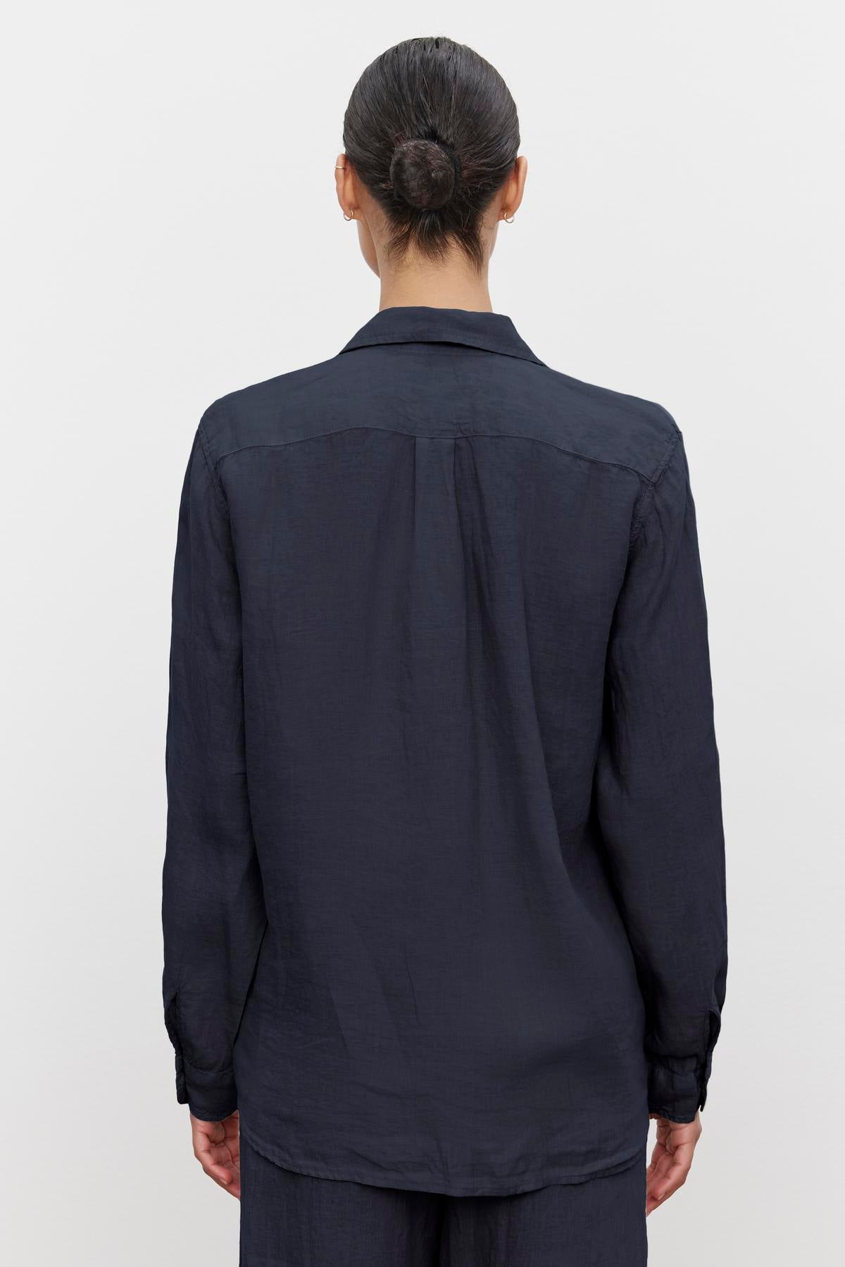 Person with dark hair tied in a bun, wearing the MULHOLLAND LINEN SHIRT by Velvet by Jenny Graham, a long-sleeve navy blue linen button-up shirt with a relaxed silhouette and scooped hemline, viewed from the back against a plain white background.-36592968499393