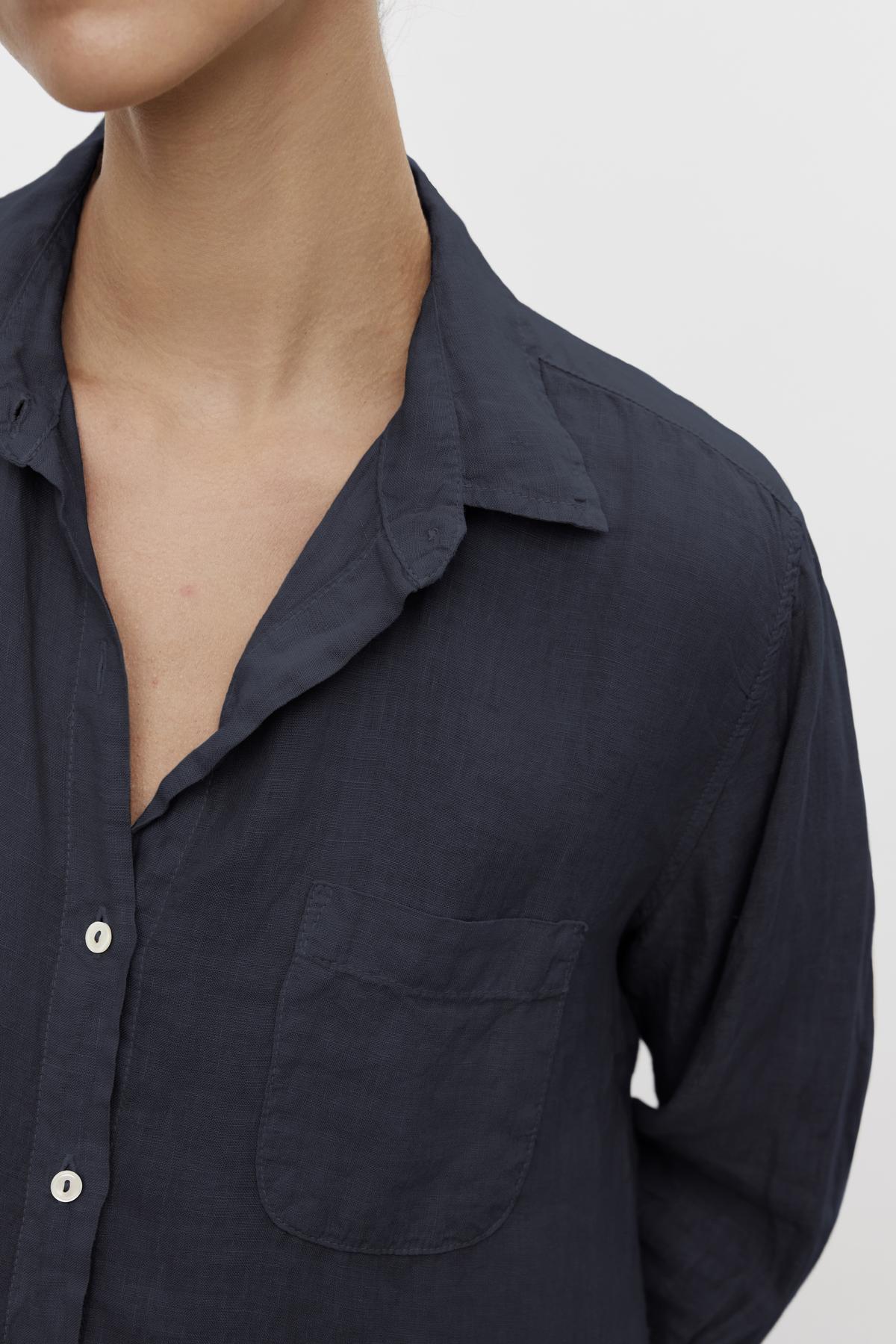  Close-up of a person wearing the MULHOLLAND LINEN SHIRT by Velvet by Jenny Graham, a dark blue linen button-up shirt with a left chest pocket. The shirt collar is slightly open, the sleeves are long, and it features a relaxed silhouette. 