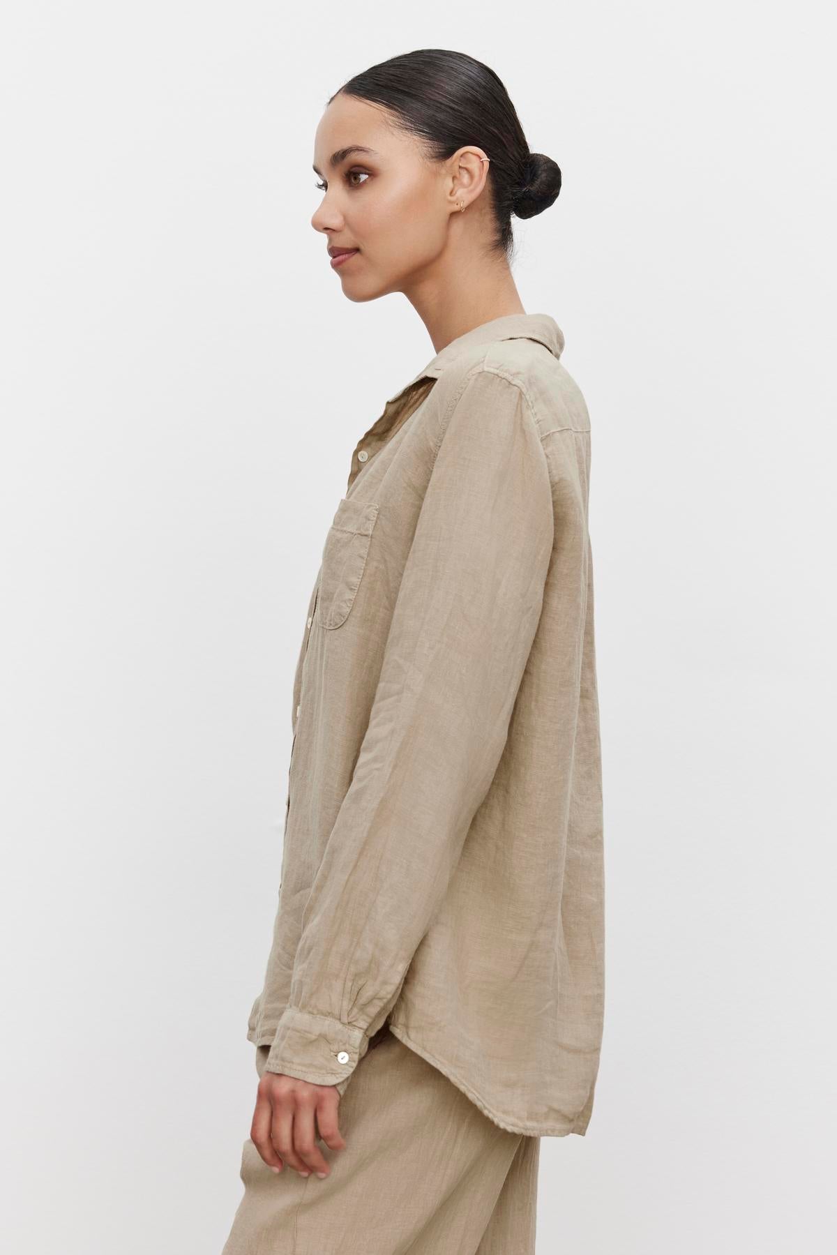 A person with dark hair tied in a bun is standing sideways, wearing a light beige linen button-up shirt and matching pants, featuring a scooped hemline against a plain white background. The shirt is the MULHOLLAND LINEN SHIRT by Velvet by Jenny Graham.-36463469691073