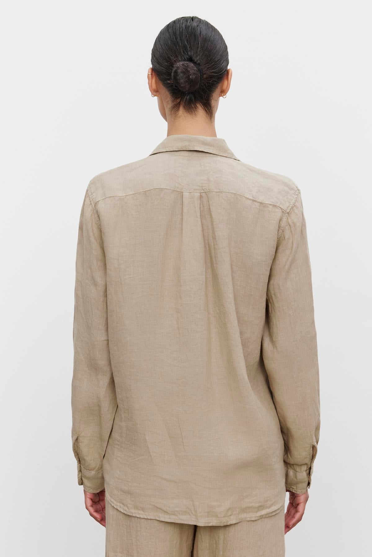   A person with hair in a bun is seen from the back, wearing a beige, long-sleeved MULHOLLAND LINEN SHIRT by Velvet by Jenny Graham with a relaxed silhouette, standing against a plain white background. 