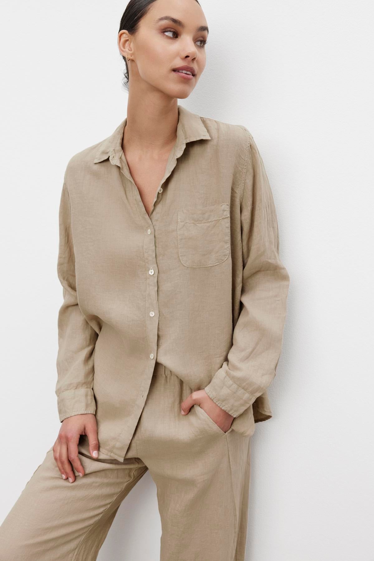   A person wearing a light brown Velvet by Jenny Graham MULHOLLAND LINEN SHIRT with a scooped hemline and matching trousers stands against a white background, with one hand in their pocket. 