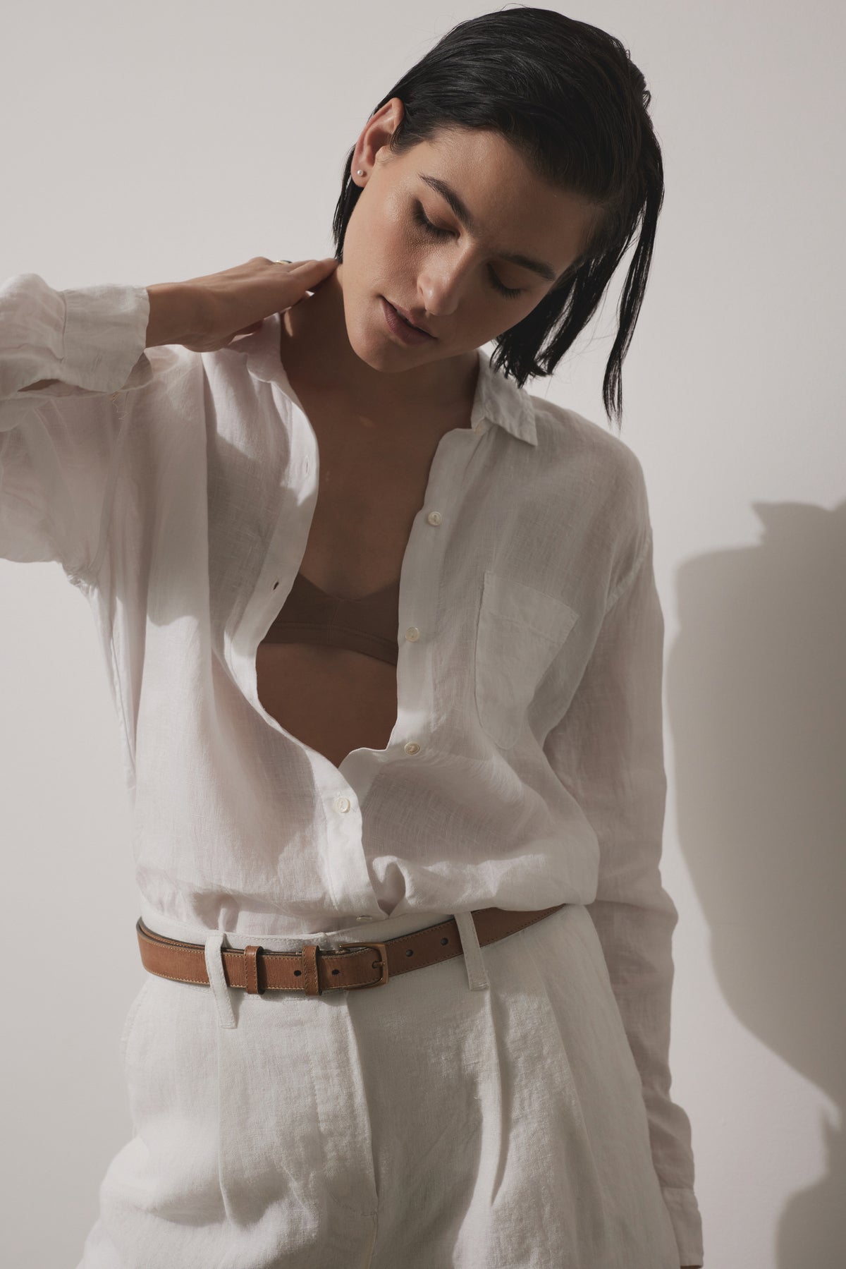 Person standing against a plain background, wearing an open white MULHOLLAND LINEN SHIRT by Velvet by Jenny Graham with a relaxed silhouette, light-colored trousers, and a brown belt, with one hand resting on the back of their neck.-37018505085121