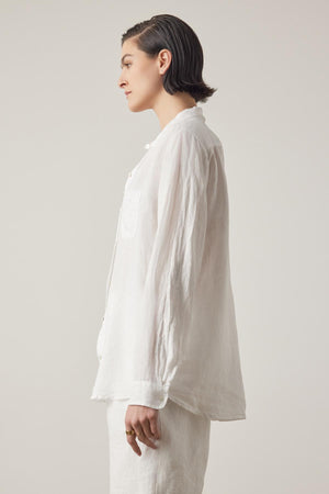 A person with short hair is standing in profile, wearing a loose-fitting, long-sleeved white MULHOLLAND LINEN SHIRT by Velvet by Jenny Graham with a relaxed silhouette and white pants against a plain background.