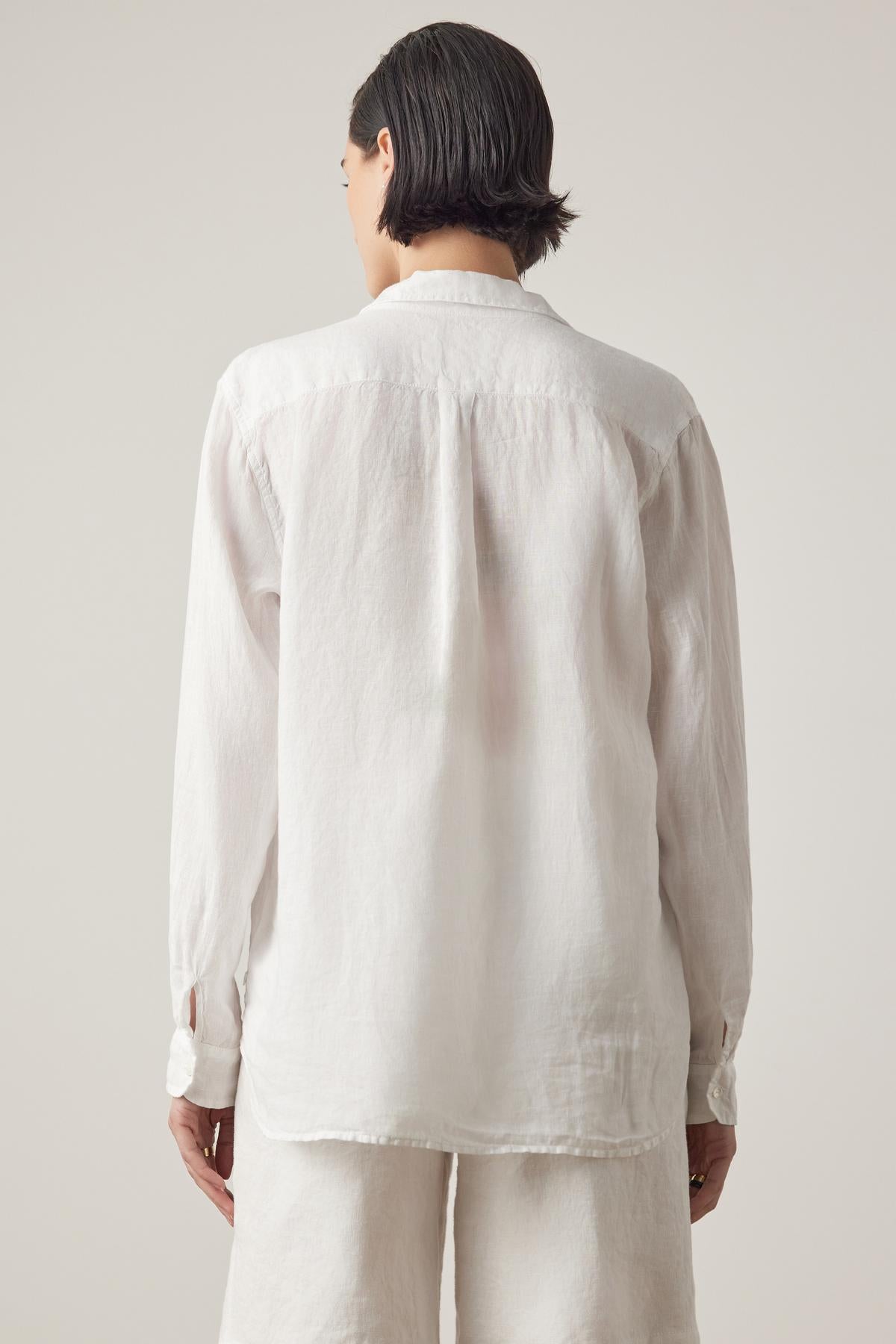 A person with short dark hair, seen from behind, wearing a long-sleeve white MULHOLLAND LINEN SHIRT by Velvet by Jenny Graham with a scooped hemline and white pants, against a plain background.-37018505183425