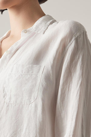 Close-up of a person wearing the Velvet by Jenny Graham MULHOLLAND LINEN SHIRT with a pocket on the left side. The focus is on the upper half of the shirt, showcasing its texture and buttons, complemented by a relaxed silhouette. The background is plain beige.