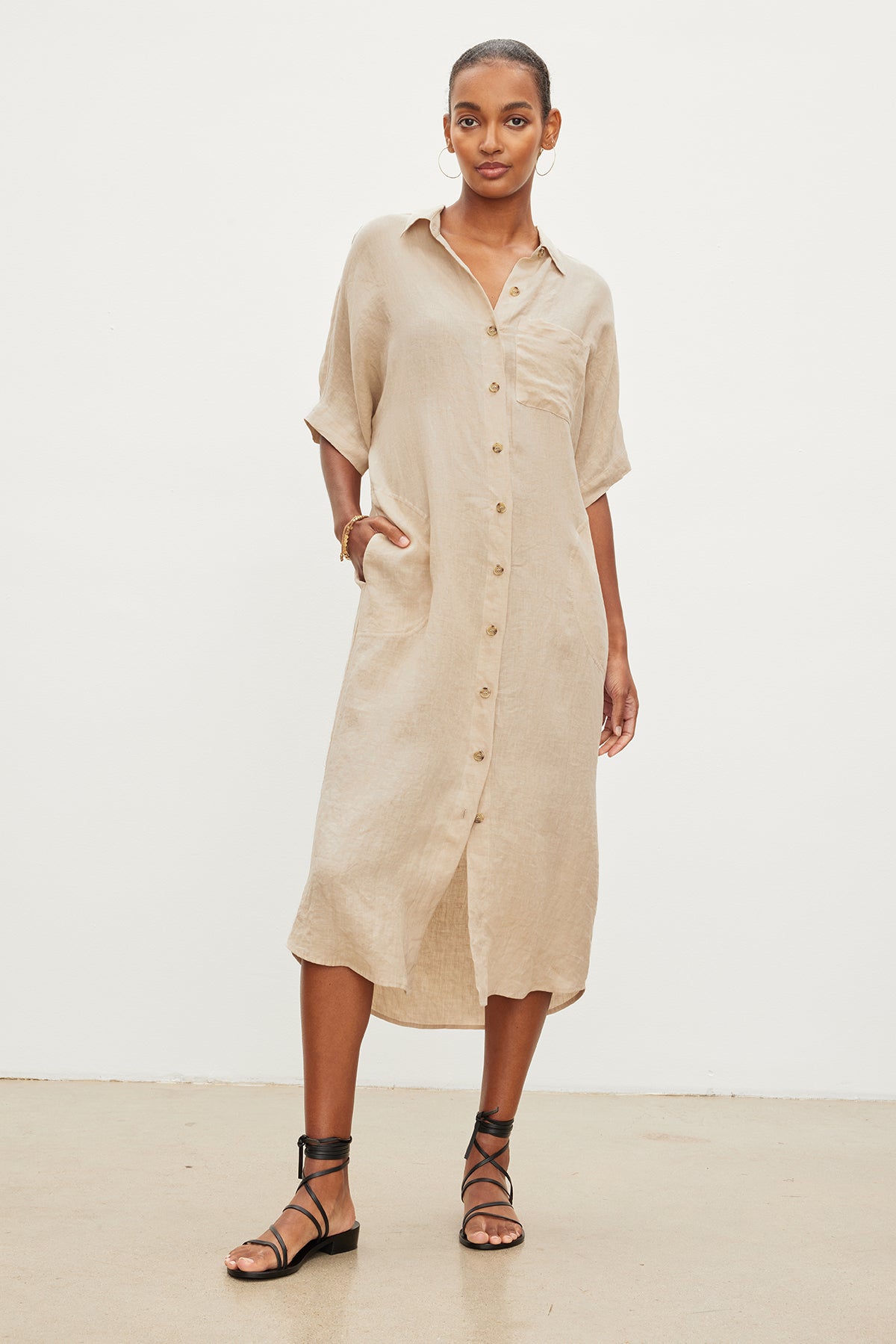   A woman in a SANDRA LINEN BUTTON-UP DRESS by Velvet by Graham & Spencer with short sleeves and buttons standing in a neutral studio setting, paired with black strappy sandals. 