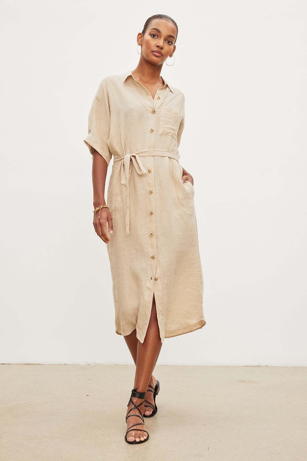 A woman models a beige mid-length SANDRA LINEN BUTTON-UP DRESS with a detachable belt, paired with black strappy sandals, in a plain studio setting by Velvet by Graham & Spencer.-35967717376193