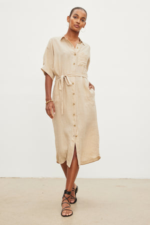 A woman models a beige mid-length SANDRA LINEN BUTTON-UP DRESS with a detachable belt, paired with black strappy sandals, in a plain studio setting by Velvet by Graham & Spencer.