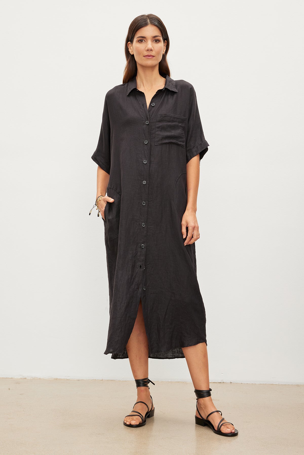   A woman standing in a studio, wearing a Velvet by Graham & Spencer SANDRA LINEN BUTTON-UP DRESS and black sandals, holding sunglasses in one hand. 