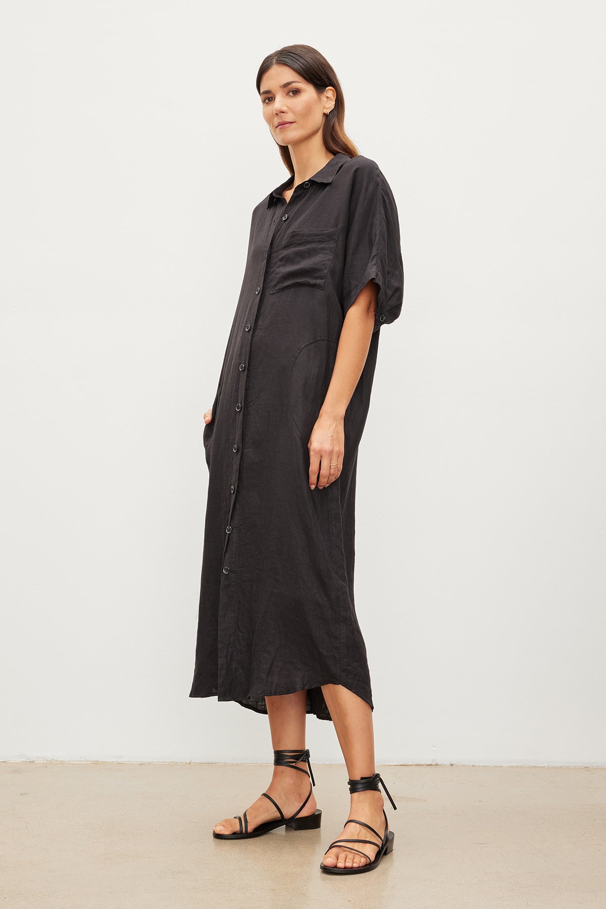 A woman stands in a plain room, wearing a black Sandra Linen button-up dress with short sleeves and strappy black sandals by Velvet by Graham & Spencer.-35967717605569