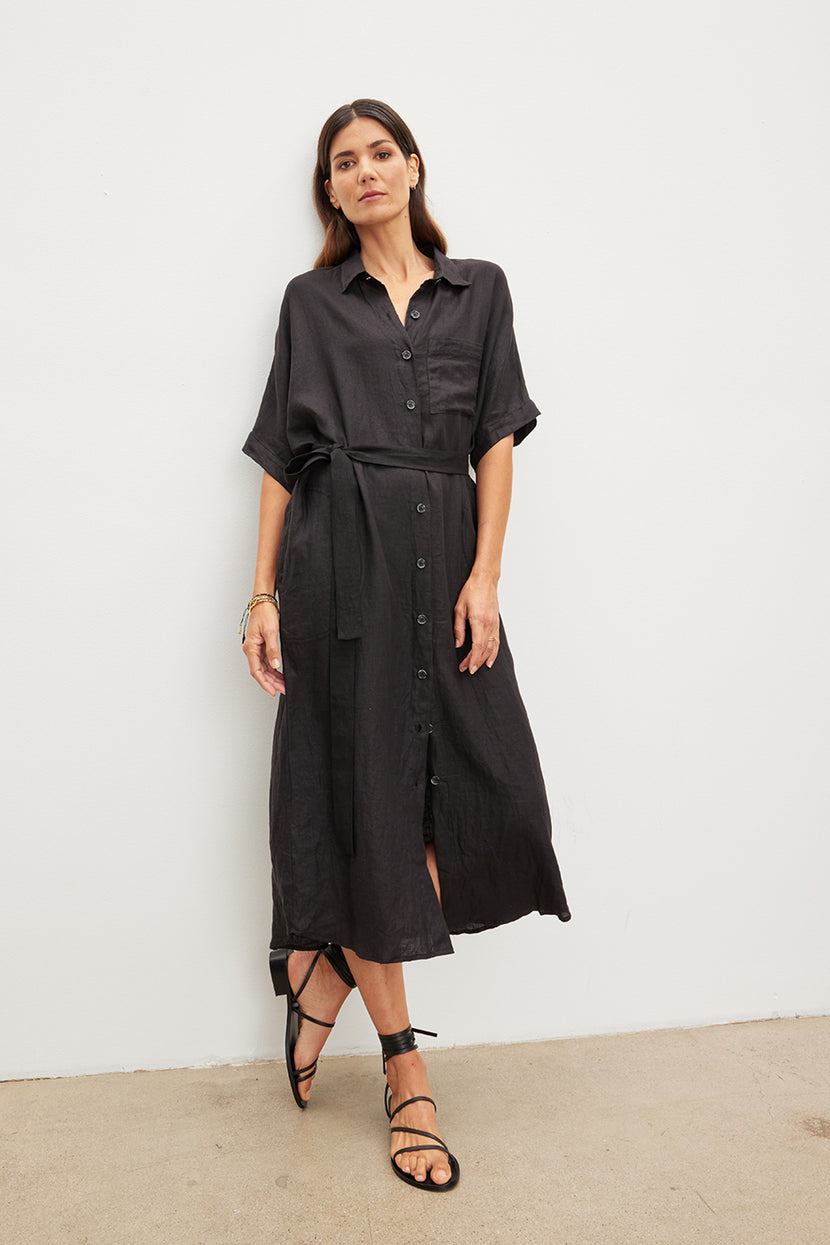 A woman stands against a white wall, wearing a black SANDRA LINEN BUTTON-UP DRESS by Velvet by Graham & Spencer with a detachable belt and black sandals.
