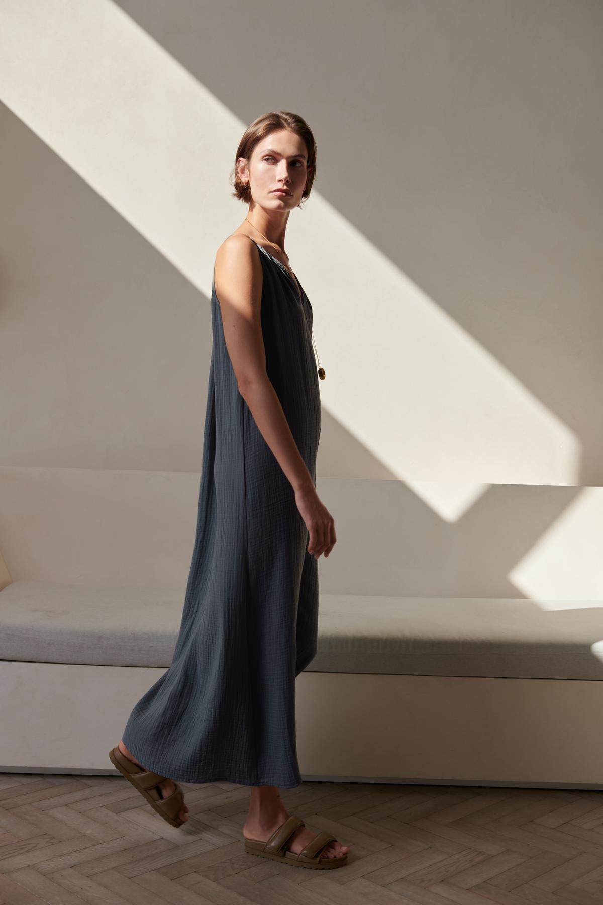 A woman in a blue cotton gauze Carrillo Dress by Velvet by Jenny Graham stands in a sunlit room with diagonal light patterns on the wall, looking thoughtfully to the side.-26293220114625