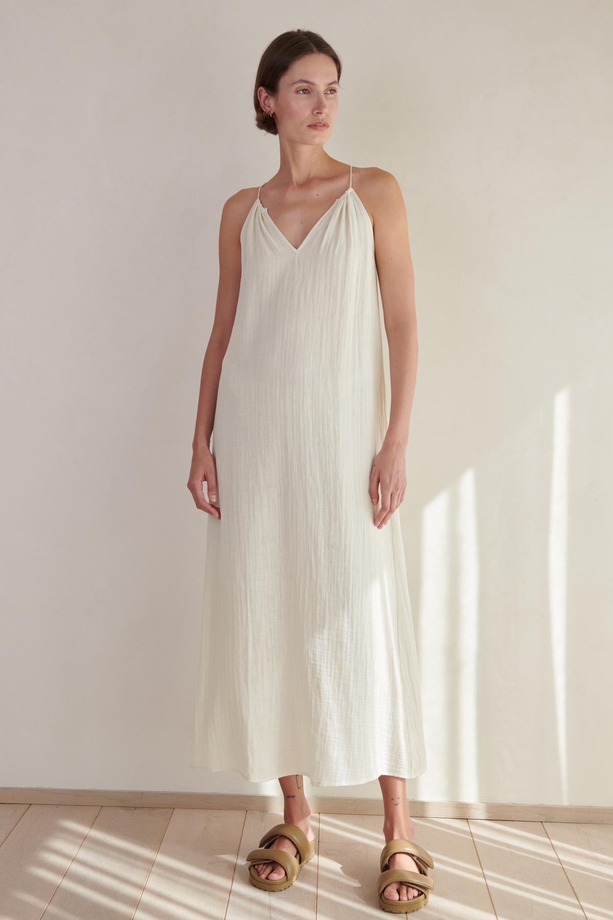 A woman in a long, white, sleeveless Carrillo dress stands in a room with soft lighting, wearing golden sandals by Velvet by Jenny Graham.-26293219754177
