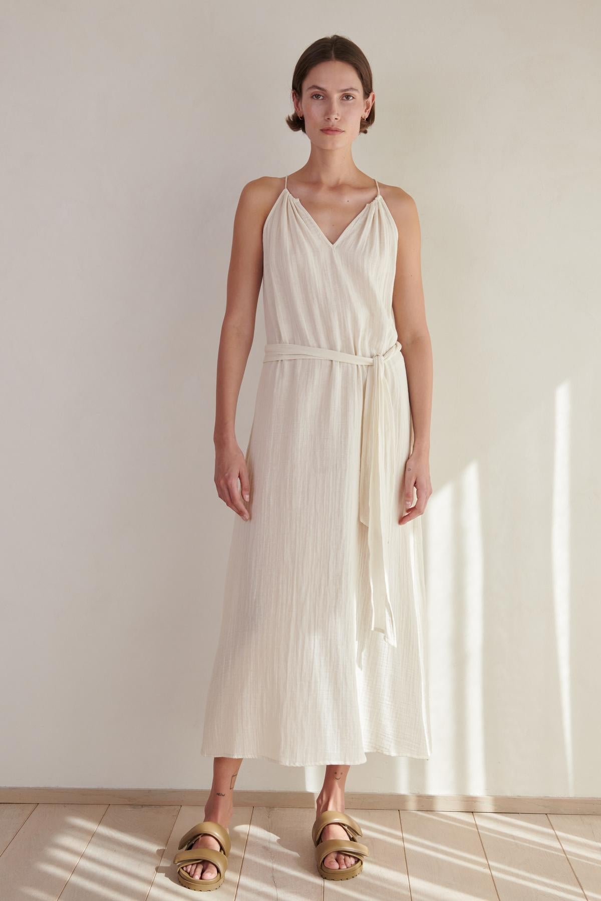 A woman in a white sleeveless CARRILLO DRESS by Velvet by Jenny Graham and gold sandals stands in a sunlit room with a neutral background.-26293219623105