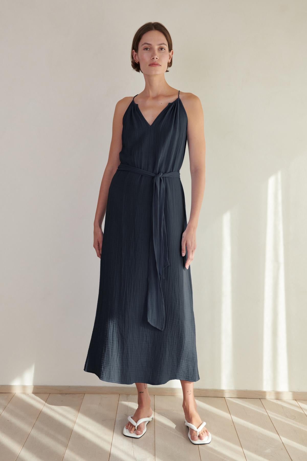 A woman in a navy blue sleeveless Carrillo dress by Velvet by Jenny Graham and white slides, standing in a softly lit room with shadows on the wall.-26293220016321