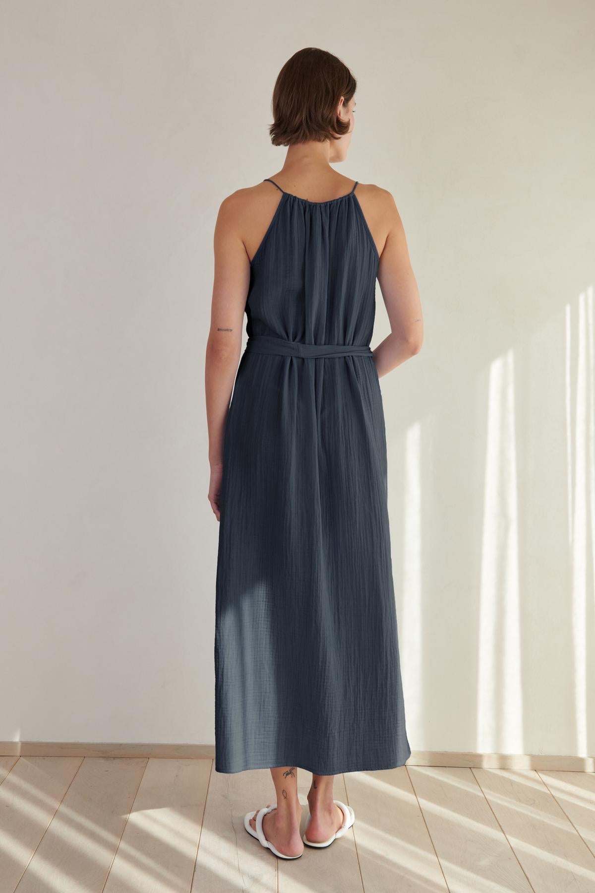 A woman stands facing away from the camera in a sunlit room, wearing a navy maxi Carrillo dress by Velvet by Jenny Graham with a halter neck and white sandals.-26293220049089
