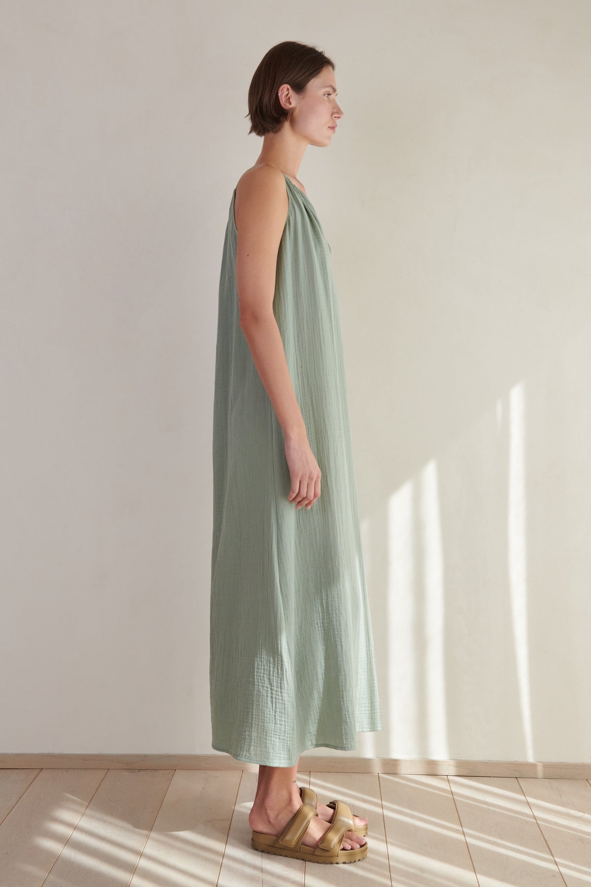   A woman stands in a sunlit room wearing a sleeveless, long green Velvet by Jenny Graham Carrillo dress with a V-neckline and beige platform sandals, looking to her left with a serene expression. 