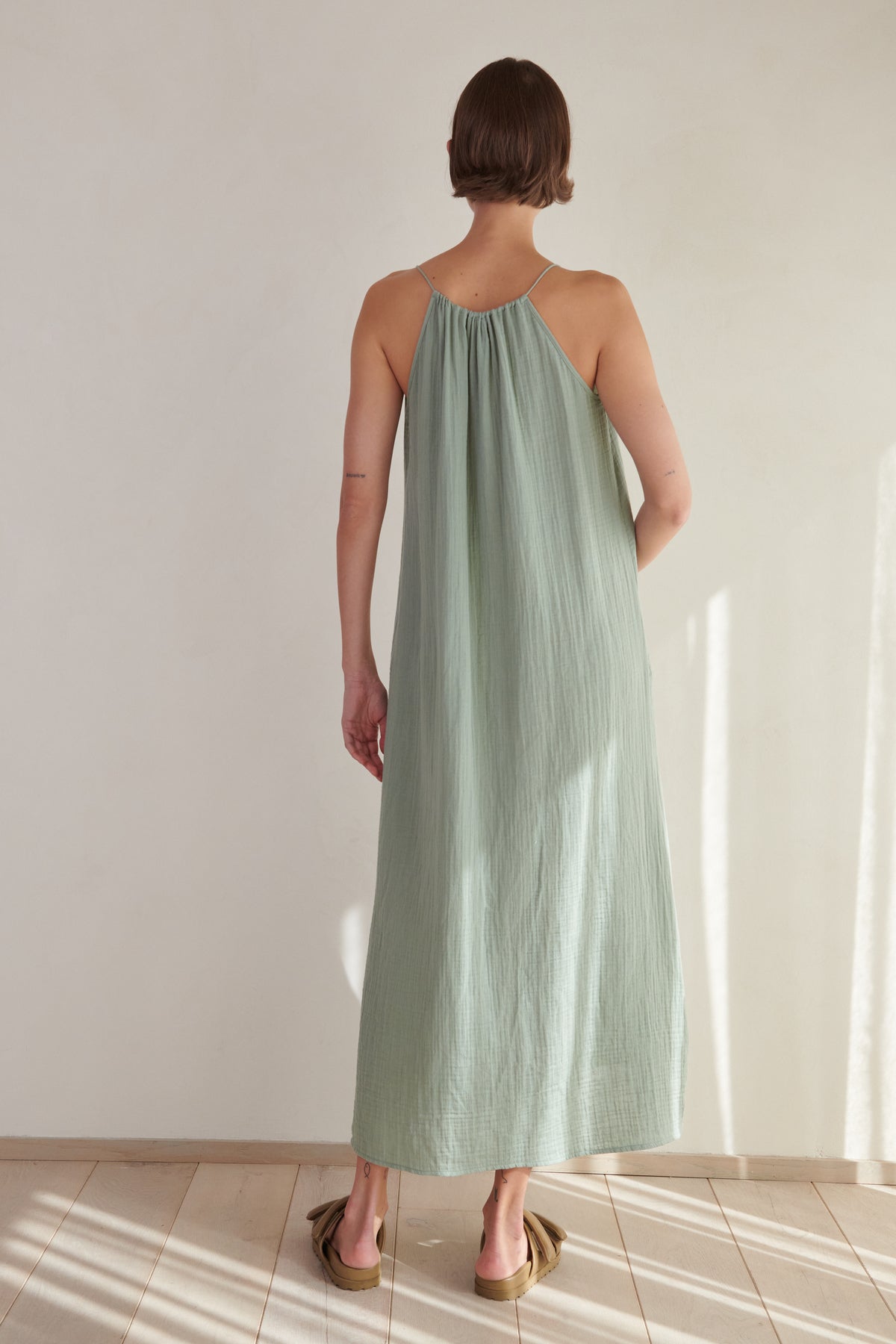 Woman standing in a sunlit room, wearing a long, light green cotton gauze Carrillo dress by Velvet by Jenny Graham with thin straps, facing away from the camera.-26293219918017