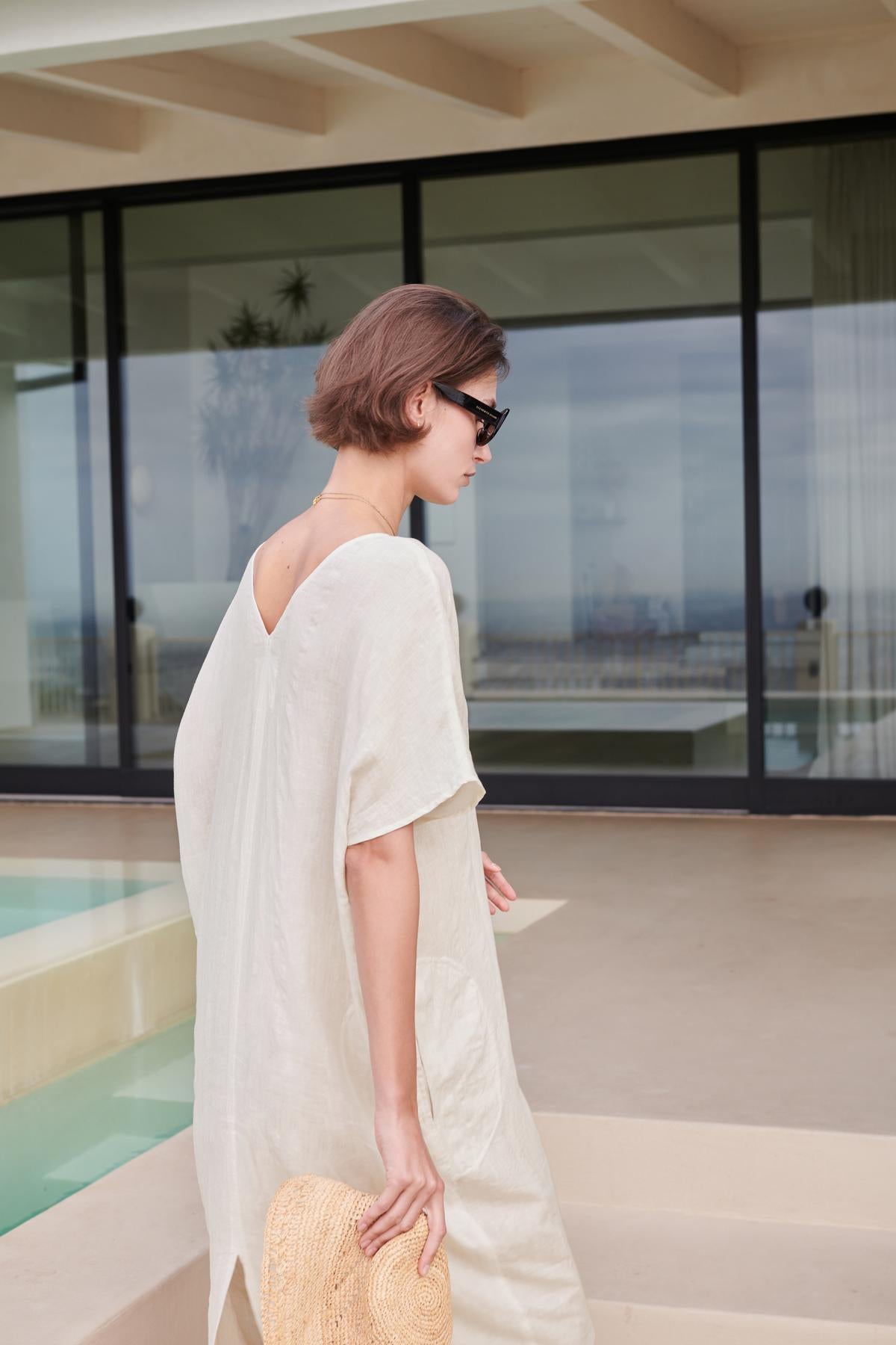 Woman in a Velvet by Jenny Graham MONTANA LINEN DRESS and sunglasses holding a straw hat, standing by a pool with a modern building in the background.-26293194817729