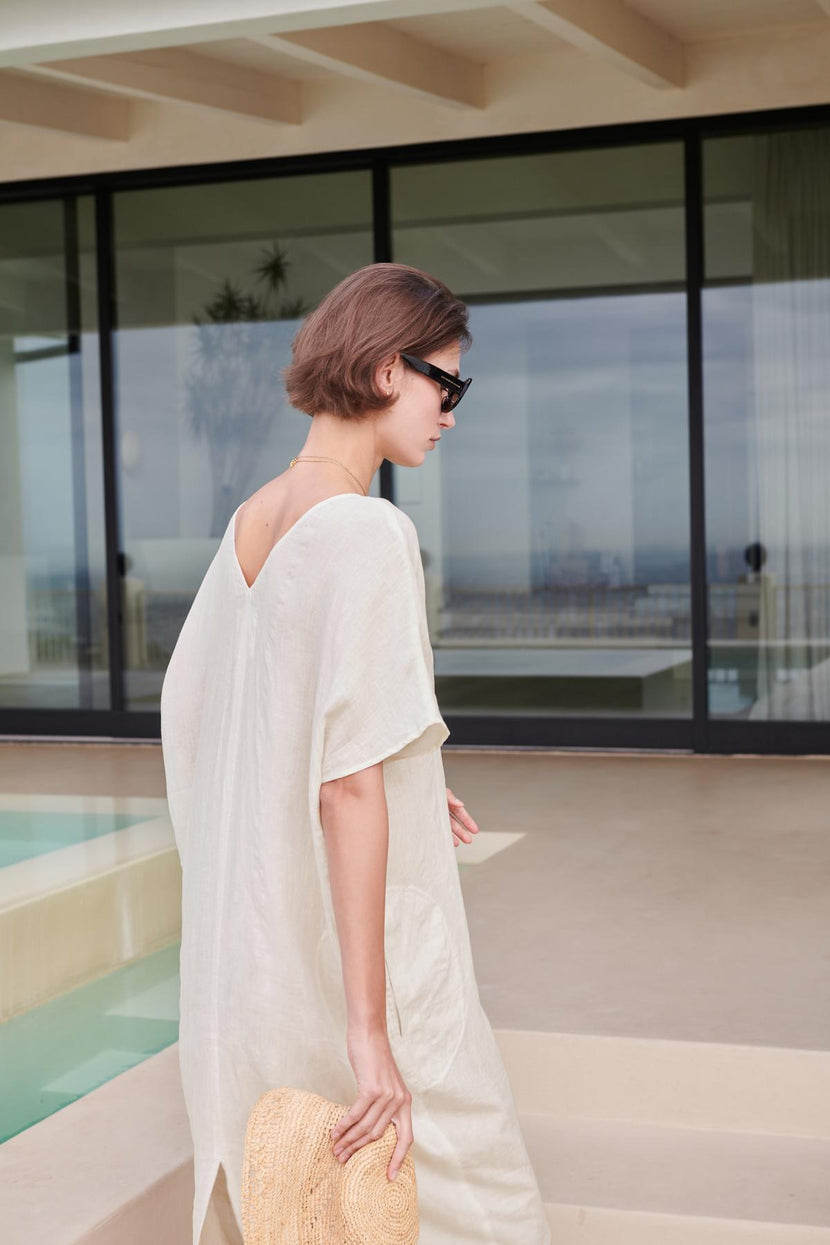 Woman in a Velvet by Jenny Graham MONTANA LINEN DRESS and sunglasses holding a straw hat, standing by a pool with a modern building in the background.