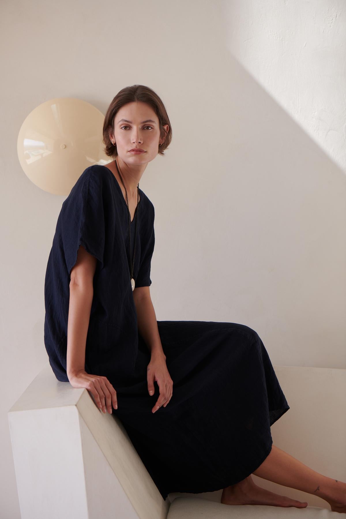 A woman in a dark blue Velvet by Jenny Graham Montana Linen Dress sitting on a white ledge beside a round wall lamp, looking at the camera with a neutral expression.-26293194588353