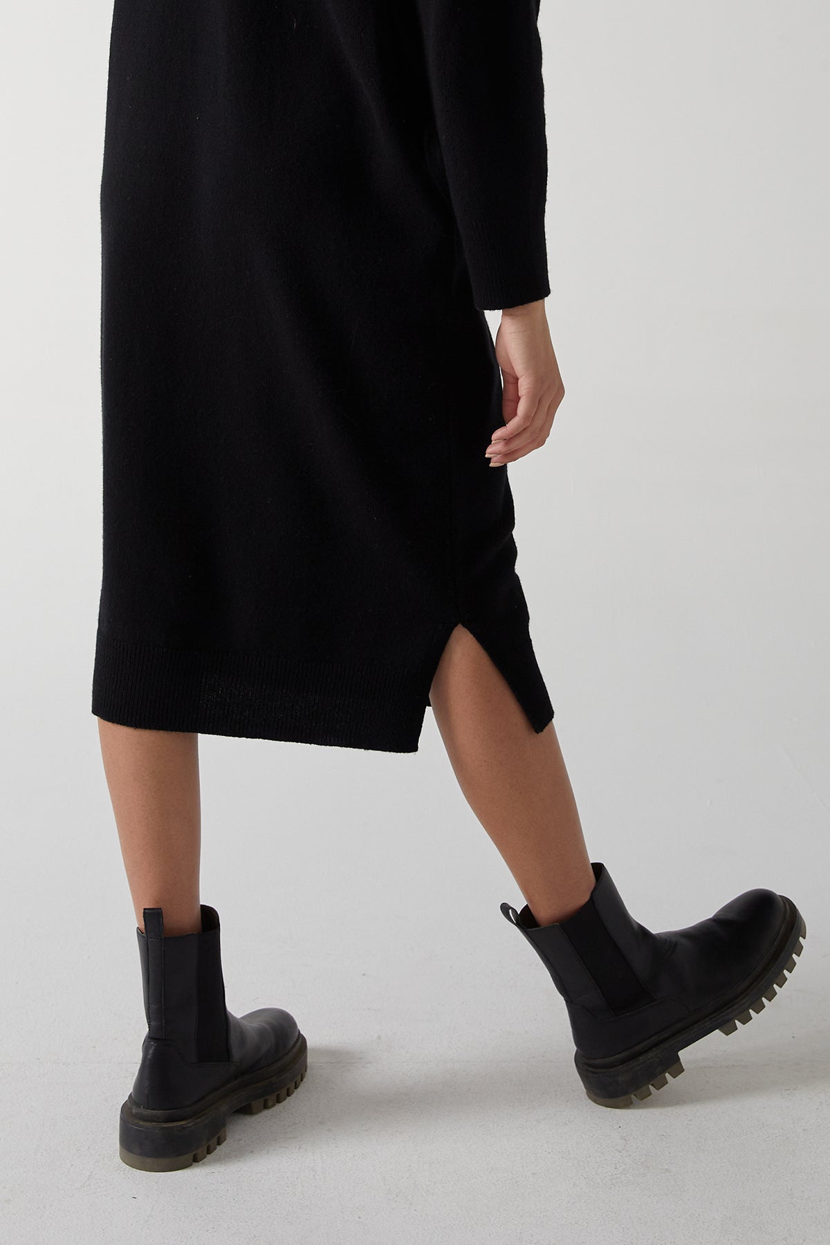 The back view of a woman wearing a Velvet by Jenny Graham LAUREL DRESS, a minimal silhouette black sweater dress with side hem slits, paired with black boots.-25315829678273