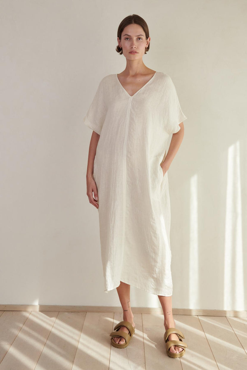 A woman wearing a long, white MONTANA LINEN DRESS with a V neckline and gold sandals stands in a softly lit room with a wooden floor. (Brand Name: Velvet by Jenny Graham)