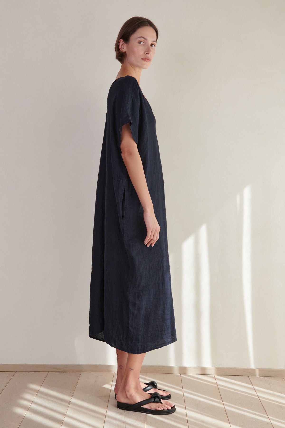 A woman in a loose navy-blue Montana linen dress by Velvet by Jenny Graham with a V neckline and black sandals standing in a room with a beige wall and tiled floor, sunlight casting shadows.-26293194653889