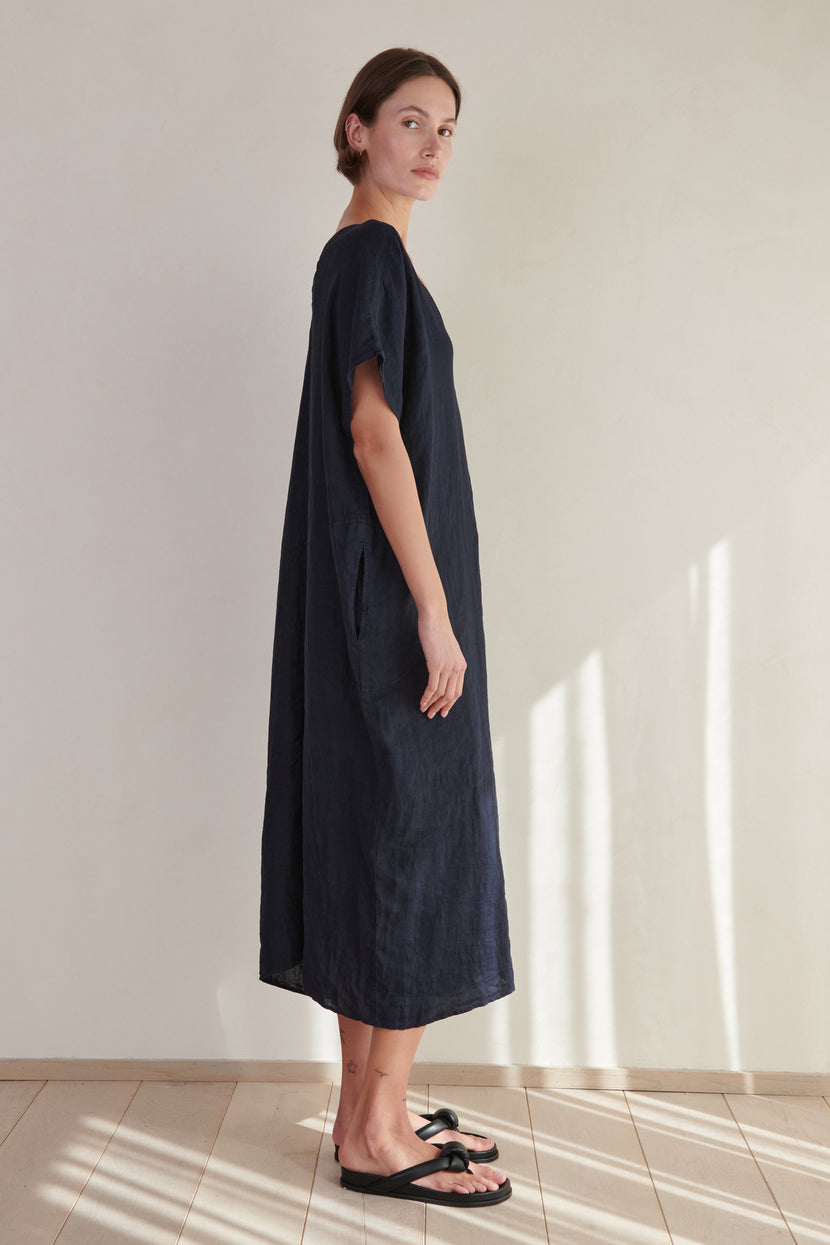 A woman in a loose navy-blue Montana linen dress by Velvet by Jenny Graham with a V neckline and black sandals standing in a room with a beige wall and tiled floor, sunlight casting shadows.