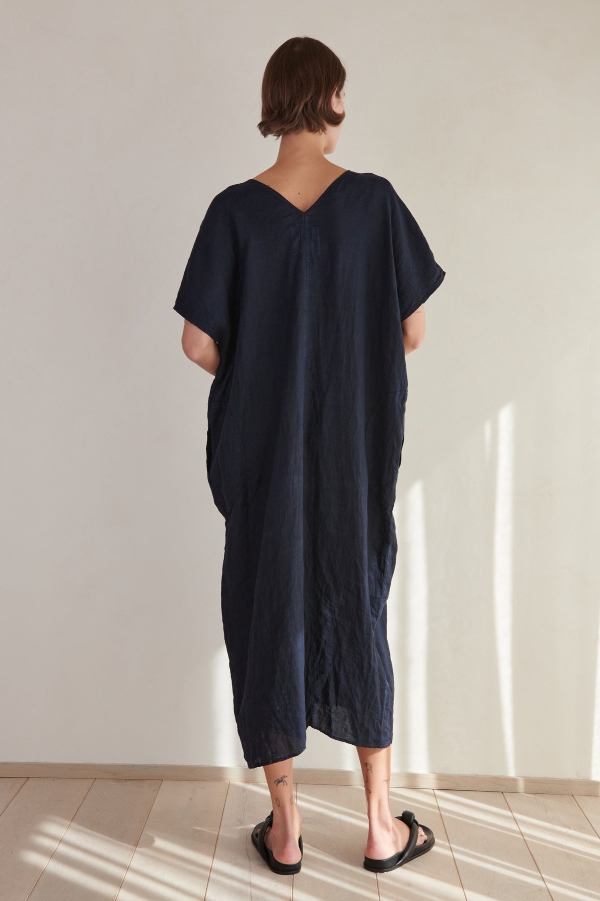A woman in a Velvet by Jenny Graham Montana Linen Dress stands facing away from the camera, highlighting a loose fit and simple design, in a well-lit room with a neutral background.-26293194686657